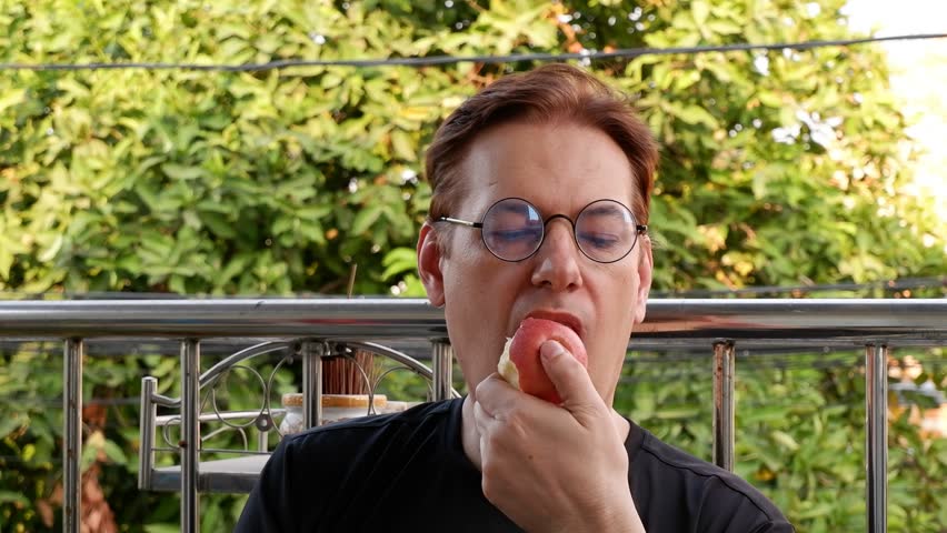 Red hair man in eyeglasses eating red apple on green trees background	