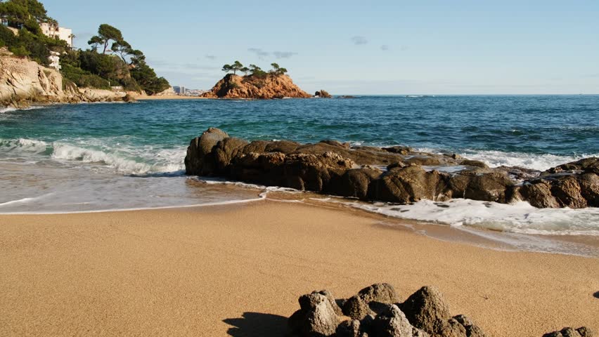 Sunny December day on Costa Brava, Spain. Waves crash against rocky shores, golden sand is covered by the tide, and the emerald sea sparkles under a clear blue sky—perfect for travel and nature visual