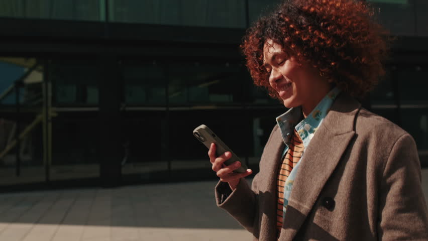 A stylish, happy woman walks down the street while looking and smiling at her phone. She is enjoying her day.