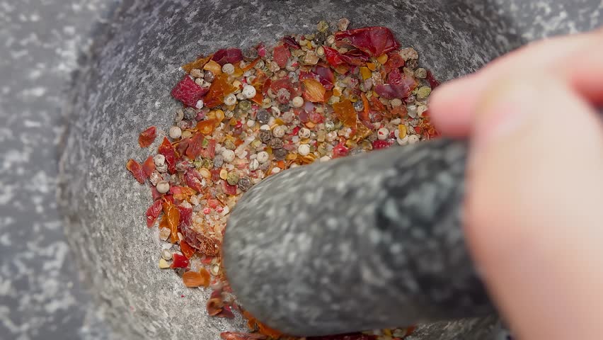 Hand with a pestle is grinding and mixing various kinds of spices, peppers and sea salt in the grey stone mortar. High quality 4k footage