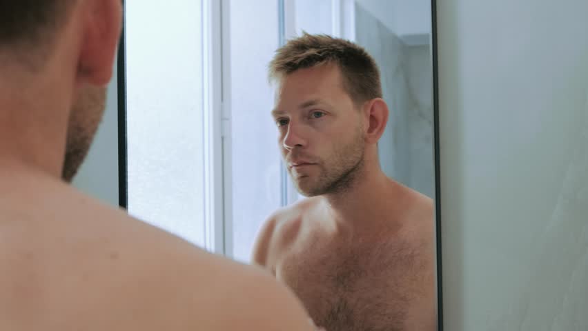 Reflection of Caucasian handsome young man applying shaving cream to face standing in the bathroom and looking at mirror. Preparation for daily morning routine male hygienic beauty procedure