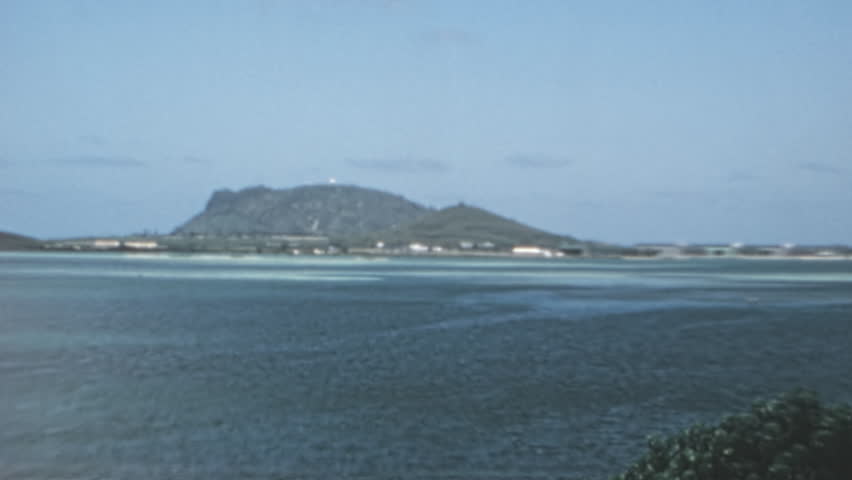 Scenic Kaneohe Bay in Oahu Hawaii with Mokolii Island rising from turquoise waters. Lush tropical vegetation frames this iconic Hawaiian landmark in a serene mid-century landscape. 1960s.