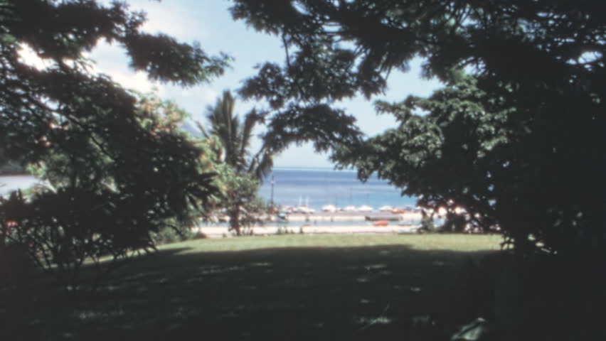 Moored yachts in Kaneohe Bay, Oahu, Hawaii, with sailboats on turquoise water framed by lush trees. Scenic vintage footage capturing Hawaiian coastal beauty and mid-century leisure. 1960s.