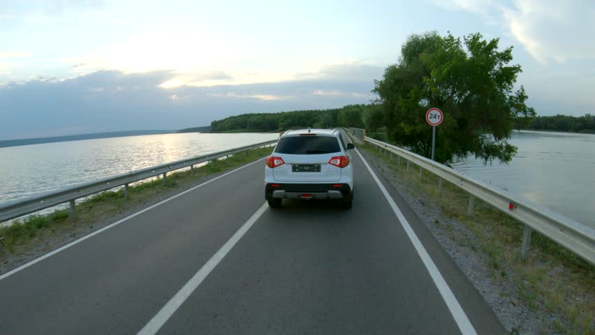 Drone follow to white car riding through dam road. SUV driving at beautiful countryside route near river or lake. Trip or journey at nature. Concept of holiday family travel. Aerial shot Slow motion