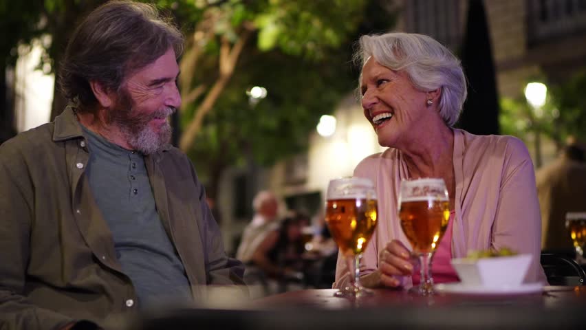 Elderly couple savoring beers, sharing intimate conversation at nighttime outdoor cafe, embodying relaxed european lifestyle and romantic connection