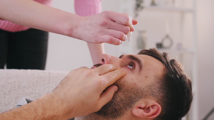 Man lies on a bed. A woman puts eyes drops in his eye.