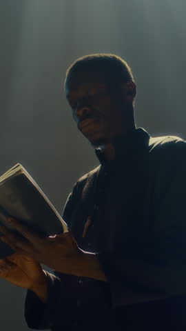 Vertical low angle view of Black pastor wearing dark cassock carefully studying bible book in catholic church