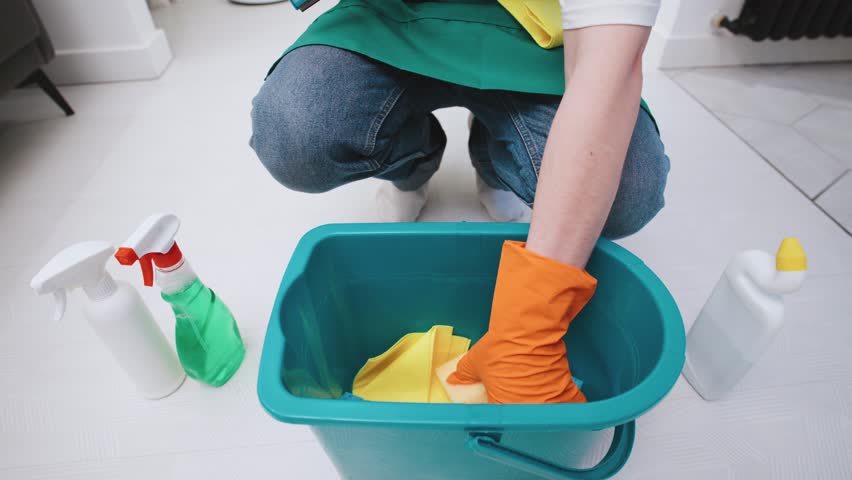 A cleaner from a cleaning service in the kitchen wearing gloves and an apron takes out cleaning products from a green cleaning bucket.