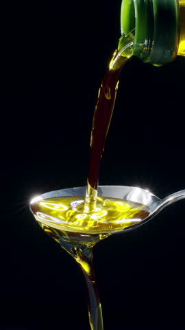 Sparkling golden olive oil is being poured into a spoon and overflowing it on a black background. Slow motion vertical template.
