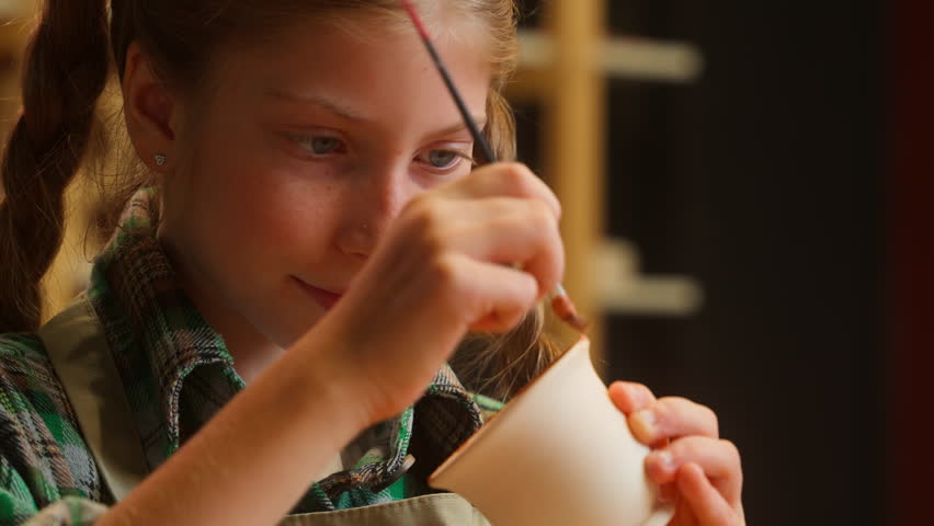 Close up cute concentrated Caucasian little girl schoolgirl child kid daughter painting clay craft drawing ornament paint brush artwork art handmade pottery mug cup ceramic crafting indoors workshop