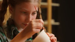 Close up cute concentrated Caucasian little girl schoolgirl child kid daughter painting clay craft drawing ornament paint brush artwork art handmade pottery mug cup ceramic crafting indoors workshop - Powered by Shutterstock - Get 15% off with code: PIKWIZARD15