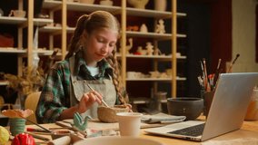 Happy smiling Caucasian teen little girl daughter child kid schoolgirl artist potter sculpting clay craft artwork pottery tool while listening online class laptop video tutorial call indoors workshop - Powered by Shutterstock - Get 15% off with code: PIKWIZARD15
