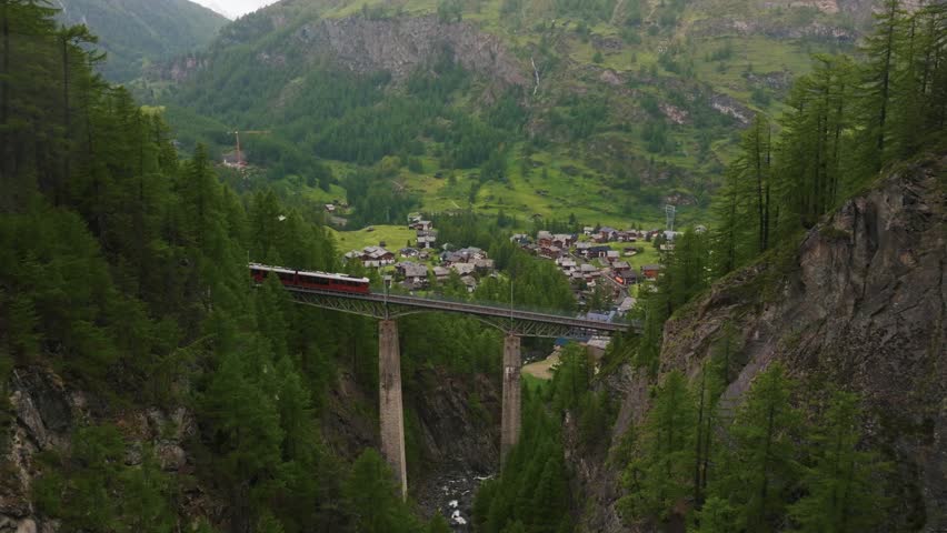 Establishing aerial shot of Gornergrat Bahn train crossing a bridge with Zermatt mountain resort in the background. Swiss Alps, Switzerland travel journey trip