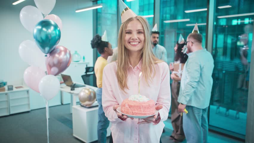 In middle of office standing birthday girl and holding cake with glowing candle. Coworkers with party hats celebrating her special day. Office room decorated with balloons and banners. Party concept.