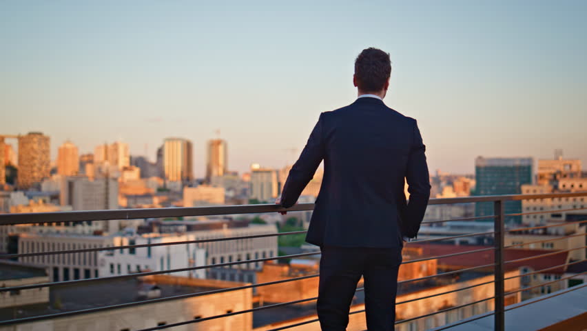Smiling ceo relaxing at outdoors terrace contemplating sunset cityscape. Happy businessman wearing elegant suit standing at balcony railings. Successful business owner looking city in evening light.