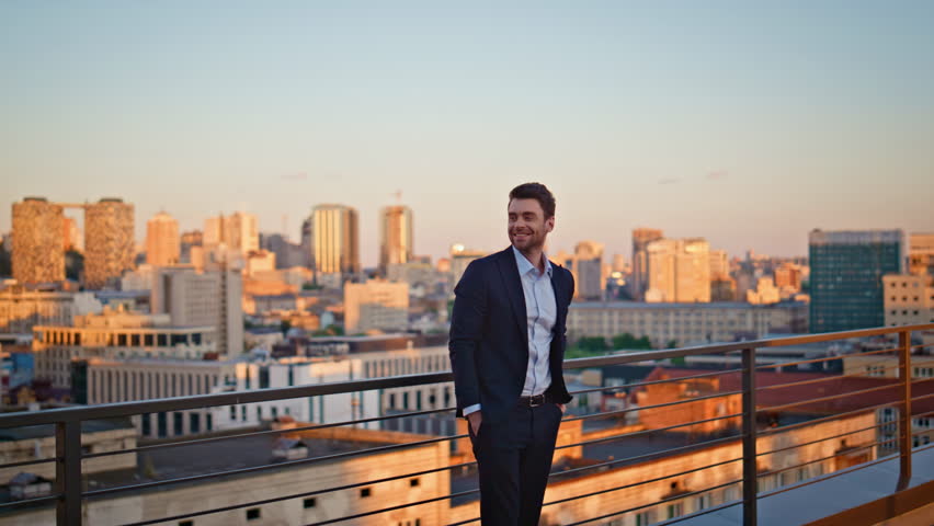 Ambitious entrepreneur standing terrace observing sunset urban view. Confident businessman relaxing on office rooftop enjoying evening cityscape. Determined man corporate leader contemplating skyline.
