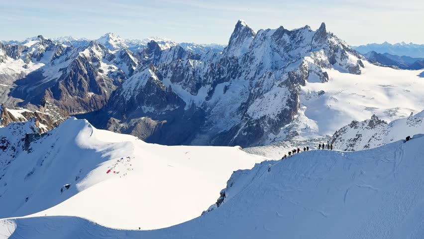 Paragliders with backpacks descend the steep snowy slopes of Aiguille du Midi, Chamonix, preparing for a parachute jump between the majestic peaks of the French Alps, with aamazing view of Mont Blanc