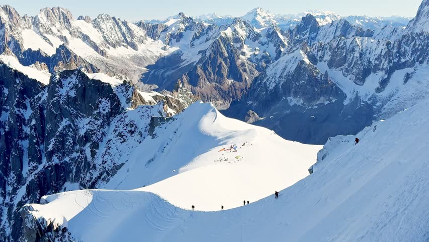 Paragliders with backpacks descend the steep snowy slopes of Aiguille du Midi, Chamonix, preparing for a parachute jump between the majestic peaks of the French Alps, with aamazing view of Mont Blanc