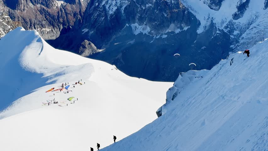 Paragliders with backpacks descend the steep snowy slopes of Aiguille du Midi, Chamonix, preparing for a parachute jump between the majestic peaks of the French Alps, with aamazing view of Mont Blanc