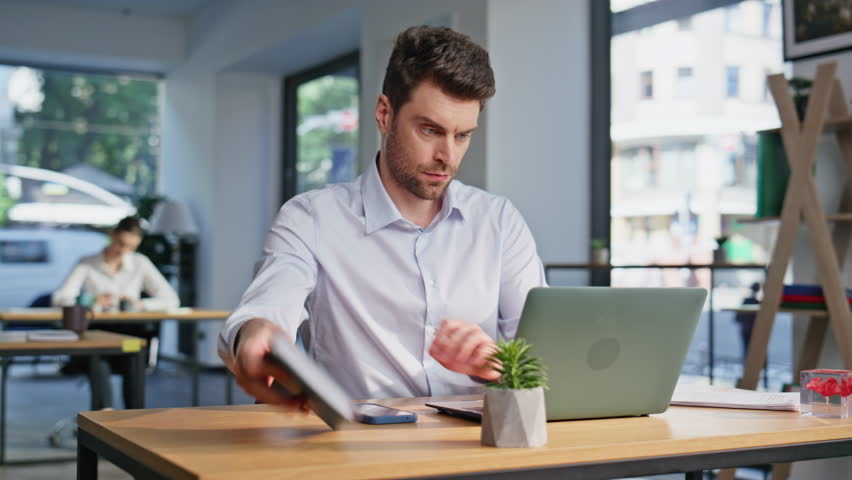 Professional manager sit down at modern workplace closeup. Focused company employee start work at contemporary coworking workspace. Serious businessman opening laptop at table in office open space.
