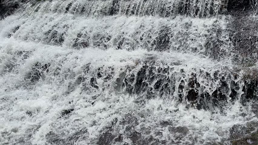 Water flows rapidly in a natural tropical river with a black rocky riverbed. Rapid river flow with black rock steps