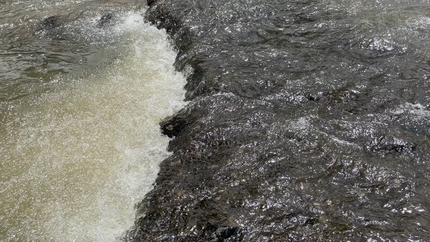 Water flows rapidly in a natural tropical river with a black rocky riverbed. Rapid river flow with black rock steps