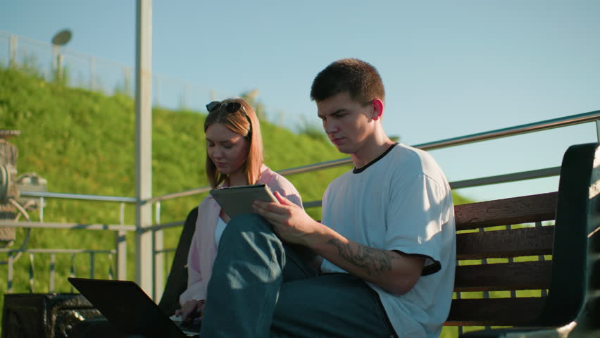 Lady typing pauses to show her friend seated close to her as he nods in agreement, both sitting on a bench outdoors with iron railings and greenery hill in the background on a sunny day