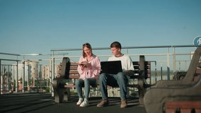 Male and female students seated outdoors on bench studying together, boy types on laptop and listens as girl reads from book, nodding in agreement, set against urban backdrop - Powered by Shutterstock - Get 15% off with code: PIKWIZARD15