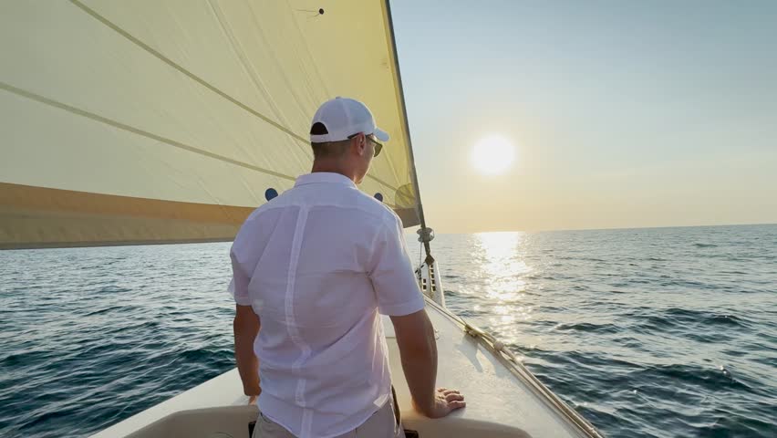 Yacht captain travels on ocean with raised hands. Man sails on yacht under white sail. Man sails on yacht under white sail. Sea voyage. Ship sails on sea, man looks at sun. Sail for dream, adventure