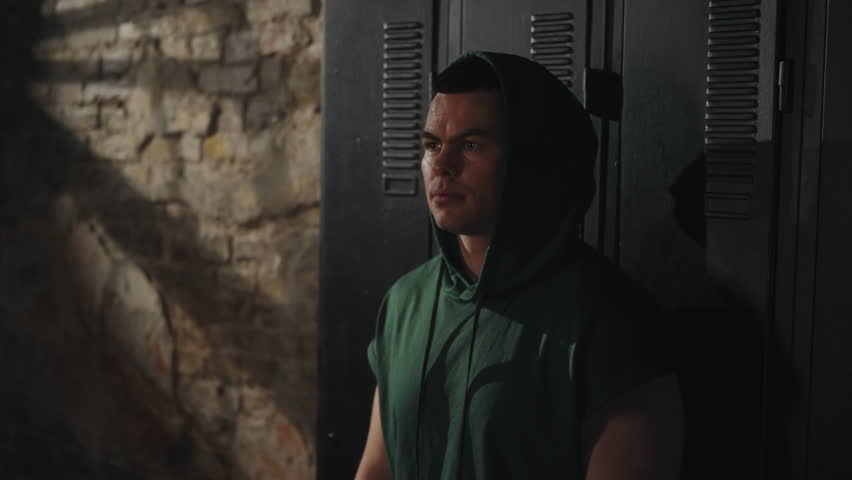 Man in hoodie leans against lockers with hand on face, wearing red hand wraps, eyes closed in exhaustion. Dim lighting. Concept fatigue and perseverance