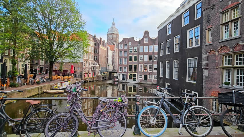 Bicycles parked on a bridge overlooking a picturesque canal in Amsterdam, surrounded by historic buildings. A large tree, boats, and outdoor cafes enhance the charm of the scenic view