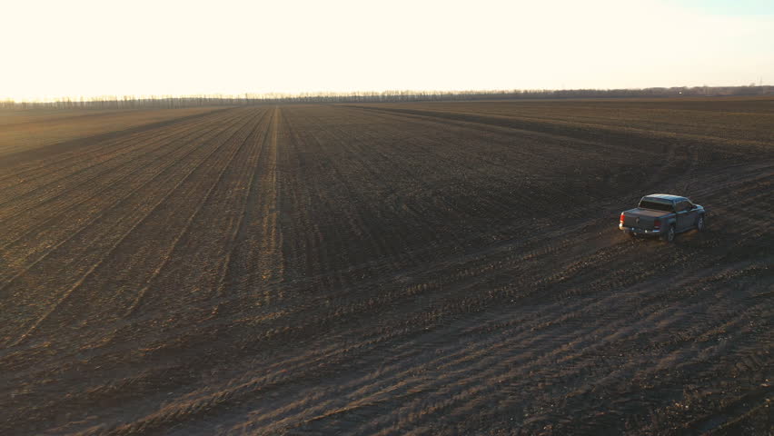 Aerial shot of pickup truck driving through plowed field after harvesting. Flying over car moving among farmland at autumn. Off road vehicle riding along ploughed meadow. Concept of agronomy farming