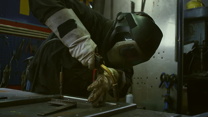 Male welder in protective gear working with metal using a welding machine producing bright sparks, flashes, and fumes, close up shot.