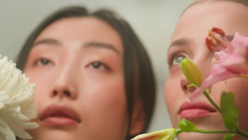 Two multiethnic diversity female faces young Caucasian girls Chinese Asian Korean Japanese women clean smooth skin natural organic cosmetics skin care shampoo make-up holding flowers perfume indoors