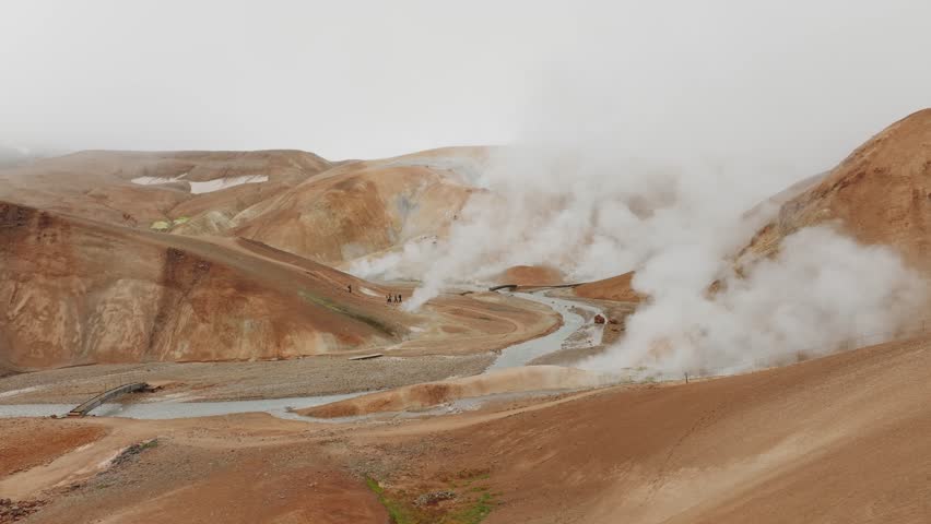50fps aerial shot of Kerlingarfjöll’s geothermal area. Steam rises from vents amidst orange hills, with visitors exploring the rugged terrain. Ideal for nature, travel, and adventure projects.