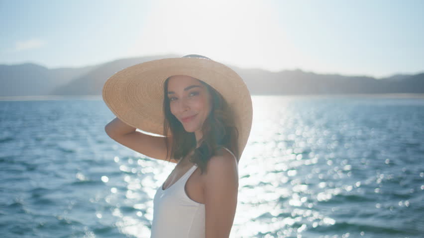 Luxurious woman relaxing on ocean shore wearing straw hat closeup. Stylish model posing summer sunlight in front bright sea water surface. Happy tourist girl standing tropical beach in white swimsuit.