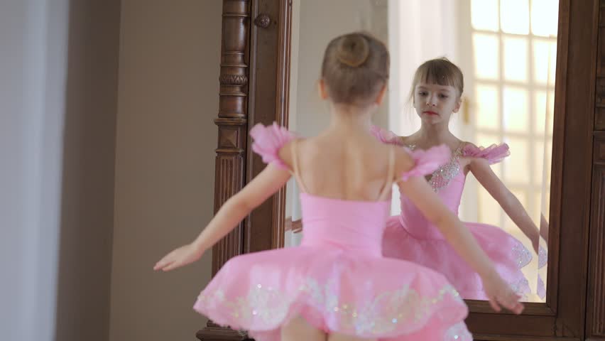 Young girl in a pink tutu is dancing in front of a mirror. Concept of innocence and joy, as the girl is dressed in a traditional ballet costume and he is enjoying herself