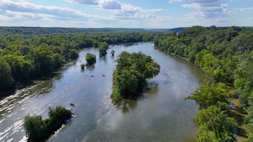 A cinematic drone shot swirls around the Potomac River on a bright sunny day. The water’s edges curve gracefully, with trees guiding the flow, creating a stunning, serene natural landscape.