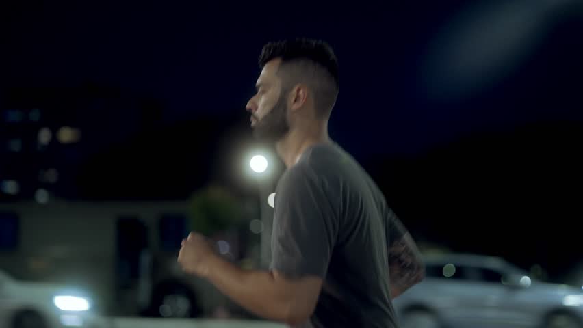 Focused man jogging through the city at night, staying determined amidst urban traffic. Athletic runner in grey t-shirt keeping a steady pace under bright city lights.