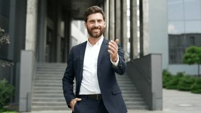 Happy handsome businessman in formal suit extending his hand to say hello while standing outside on the street near an office building. Friendly smiling entrepreneur or worker greets looking at camera - Powered by Shutterstock - Get 15% off with code: PIKWIZARD15