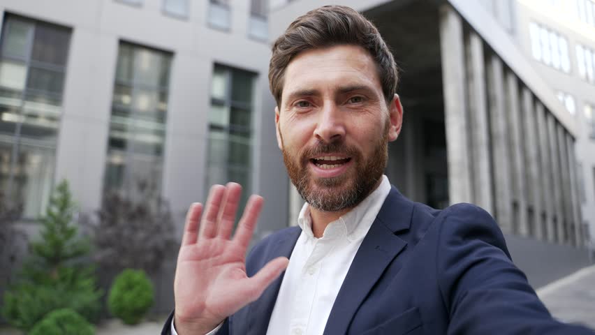 POV. Smiling businessman in a formal suit talking on a video call using a mobile phone standing on the street near an office building. Entrepreneur communicates remotely looking at camera. Online chat - Powered by Shutterstock - Get 15% off with code: PIKWIZARD15