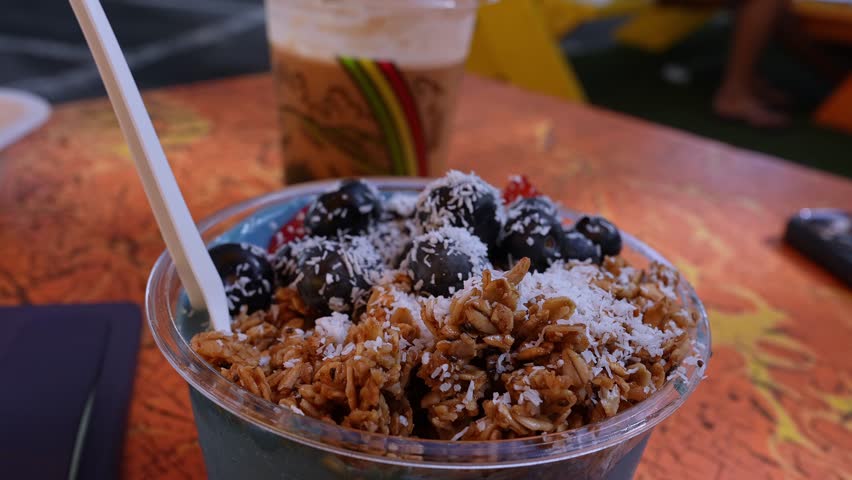 A vibrant acai bowl with granola, coconut, and blueberries on an orange table. Background shows iced coffee, a hand with food, and a parked car.