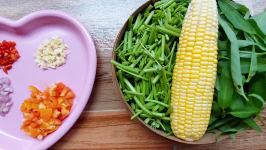 A heart-shaped plate filled with small pieces of onion, garlic, red chili, and tomato. Next to it is a basket filled with corn, kale stalks, and kale leaves. Ingredients for stir-fried kale