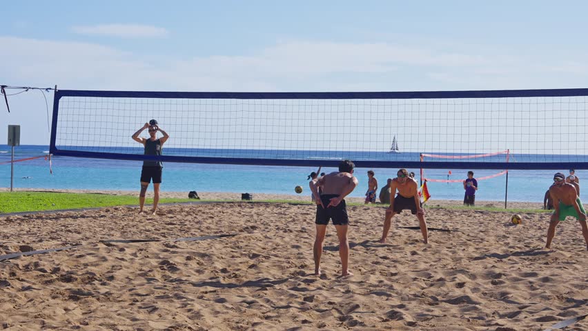 Honolulu, Hawaii. January 10, 2025. A lively beach volleyball game on a sandy shore in Hawaii. Players are mid action, with a sailboat in the distance and people relaxing nearby.
