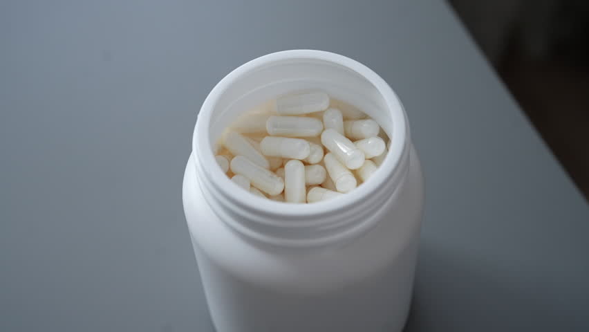 Male hand reaching for white pill from white container on gray table. Close-up of unrecognizable man taking capsule from plastic bottle. Concept of medicine, healthcare, pharmaceutical treatment.