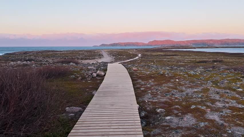 Long hiking trail made of wooden planks leads to horizon. It offers beautiful northern landscape with tundra and ocean in foreground, against backdrop of rocks and mountains, at sunset.