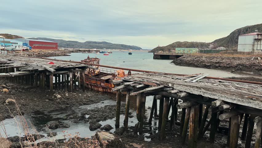 Old, abandoned wooden bridge in fishing port town was destroyed by flood from nearby river. rusted ship stranded next to bridge. Teriberka, Russia