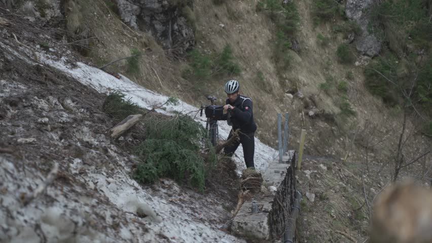 Man cyclist pulls bicycle through avalanche in mountains. Bicyclist is trapped in obstacle and is pulling his cycle through an ice slide with bicycle in his hands. Avalanche on cycling route in Alps. 