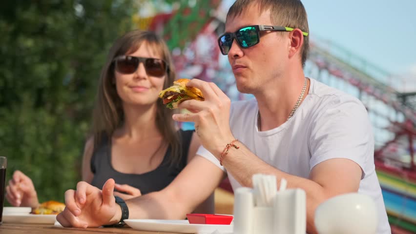 A couple sits at a summer restaurant table, smiling and enjoying their weekend at an amusement park. The man eagerly eats a burger while the woman, wearing sunglasses, keeps the conversation flowing.