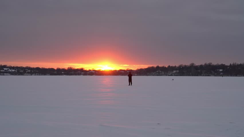 Breathtaking winter sunset casts a warm glow over a vast frozen lake with a solitary figure standing in the distance